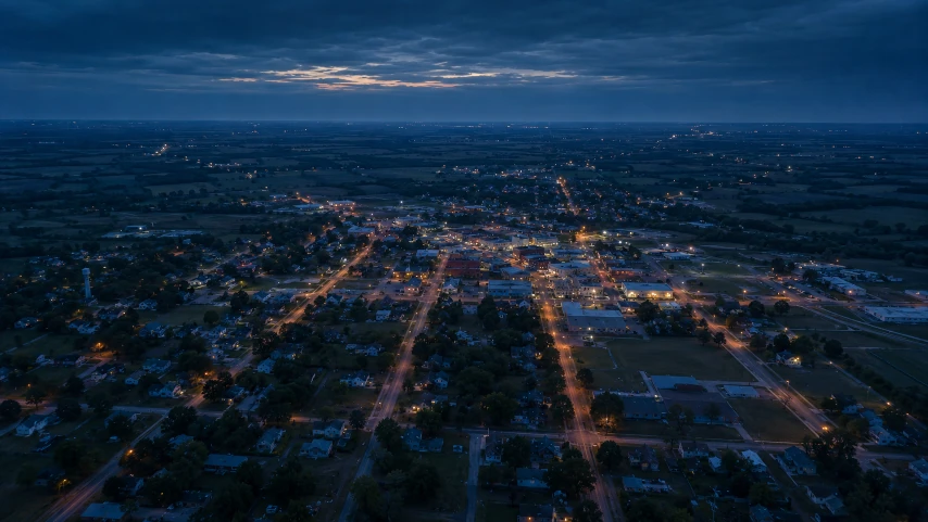 Aerial view of a small American town at dusk, amber streetlights tracing the grid against dark fields